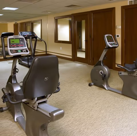 Indoor exercise room with stationary recumbent bikes and other cardio equipment, beige carpeted floor, wood-paneled doors, and beige walls with windows looking into adjacent rooms.