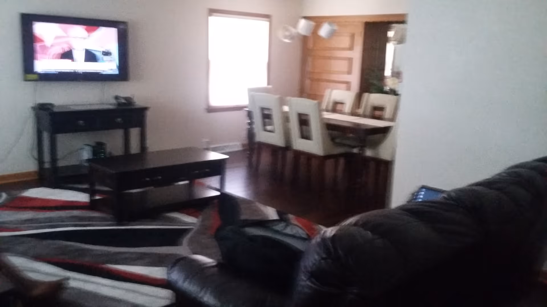 Interior view of a living room and dining area. The living room has a dark leather couch, a patterned area rug, a black coffee table, and a TV mounted on the wall above a small black console table. The dining area features a rectangular dining table with six white upholstered chairs and a window letting in natural light.
