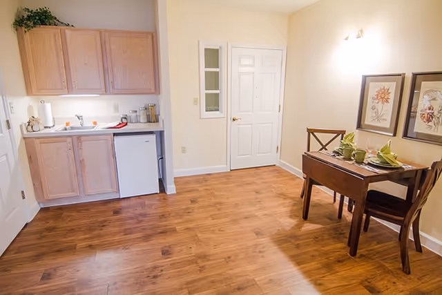 Small apartment interior showing a kitchenette with sink and cabinets on the left and a two-seat dining table on the right on hardwood floors.