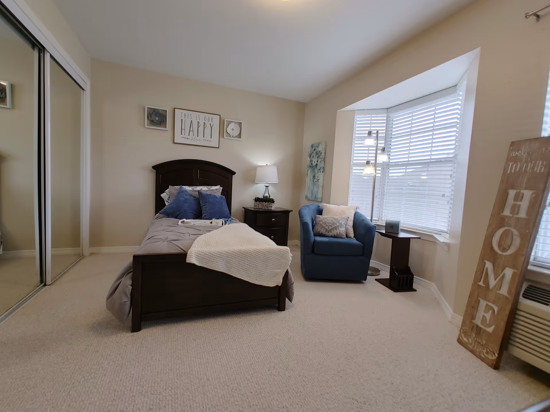 A cozy bedroom featuring a dark wooden bed with gray bedding and blue pillows, a matching nightstand with a lamp, a blue armchair with cushions near a large window with white blinds, a floor lamp, and wall decorations including a sign that reads 'This is our HAPPY home' and a tall wooden sign leaning against the wall that says 'HOME'. There is also a mirrored closet door on the left side.