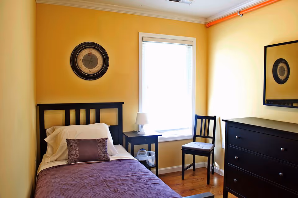 Yellow-walled bedroom with single bed, dresser, nightstand, chair, and a window.