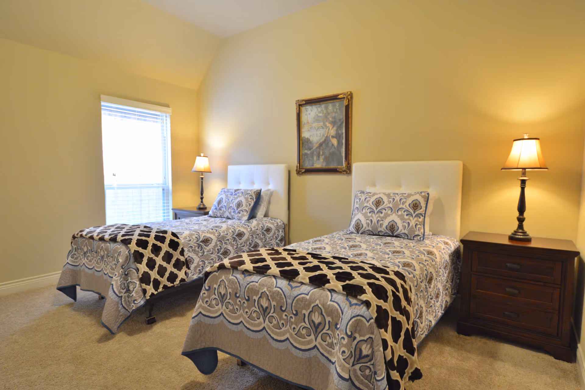 A bedroom with two twin beds featuring white tufted headboards and patterned bedspreads in shades of gray, blue, and beige. Each bed has a matching pillow and a dark brown and beige throw blanket. There are two dark wooden nightstands, each with a lamp, and a window with blinds on the left wall. A framed artwork hangs on the wall between the beds.