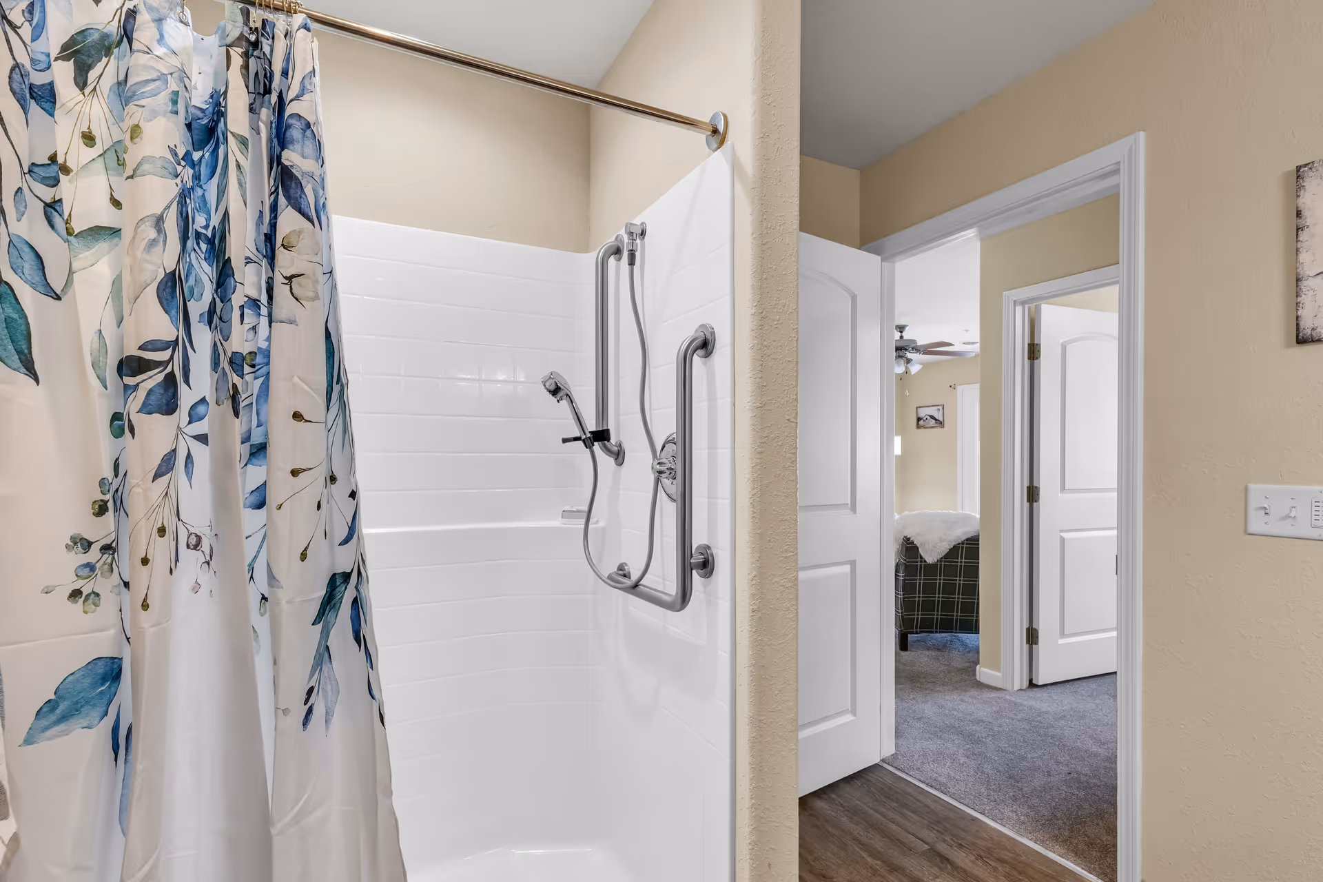 A white-tiled shower with a floral curtain and grab bars, seen next to an open doorway showing a bedroom beyond.