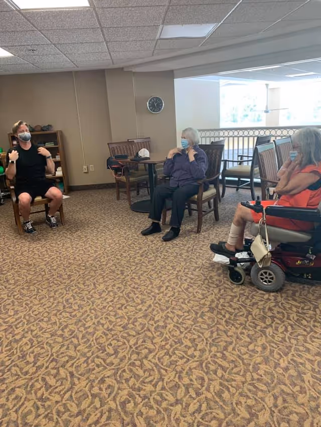 Three elderly individuals wearing face masks are seated in a common area with carpeted flooring and beige walls. One person is in a motorized wheelchair, and the other two are seated on wooden chairs. They appear to be engaged in a group activity or exercise session. A clock is visible on the wall behind them.