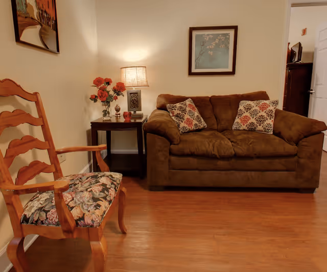 A cozy living room area with a brown cushioned sofa adorned with two patterned pillows, a wooden chair with floral upholstery, a side table with a lamp, flowers, and decorative items, and framed artwork on the walls.