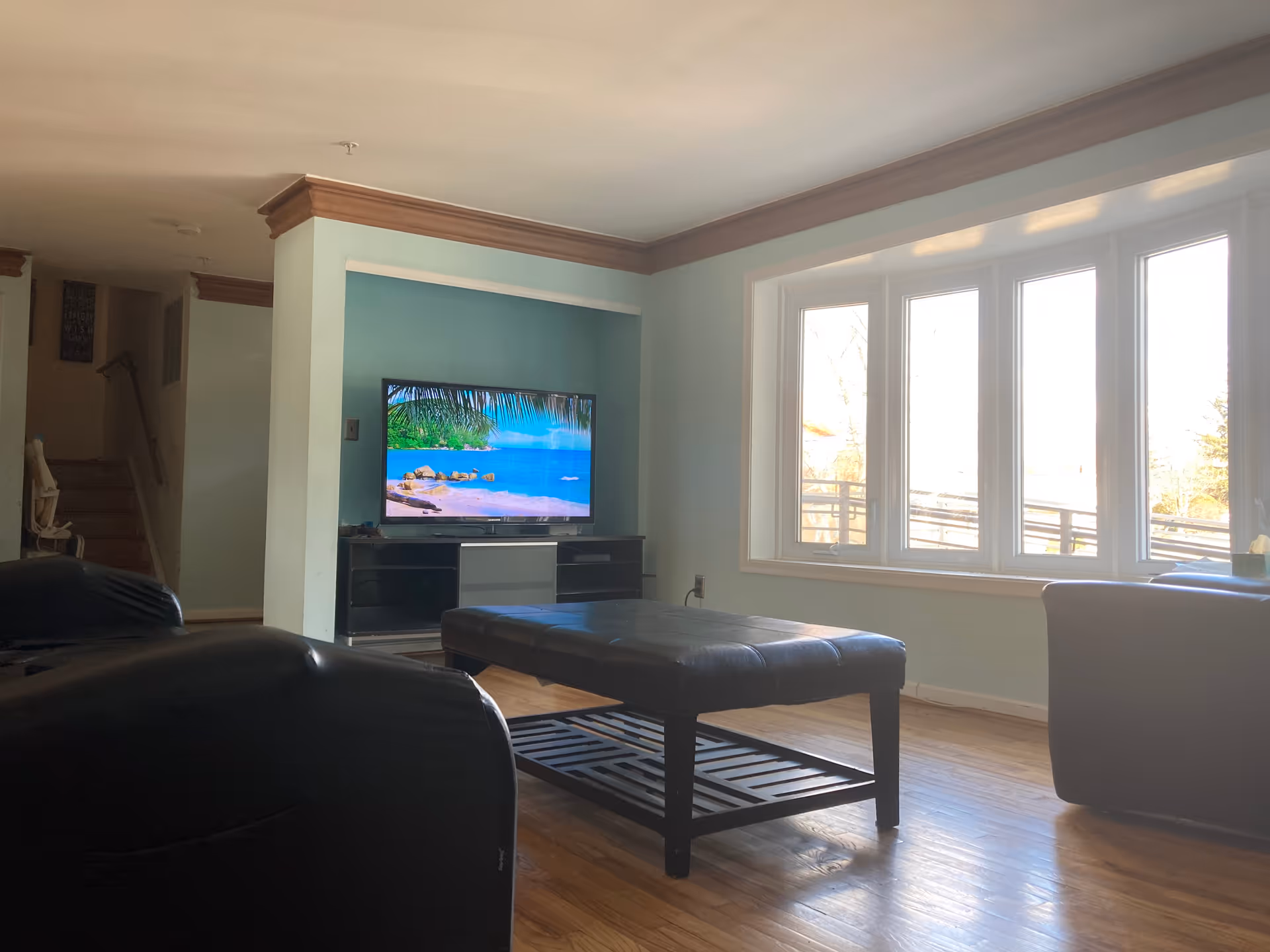 A living room with a large window letting in natural light, a flat-screen TV displaying a beach scene, a black leather ottoman coffee table, and black leather chairs. There is a staircase visible in the background.