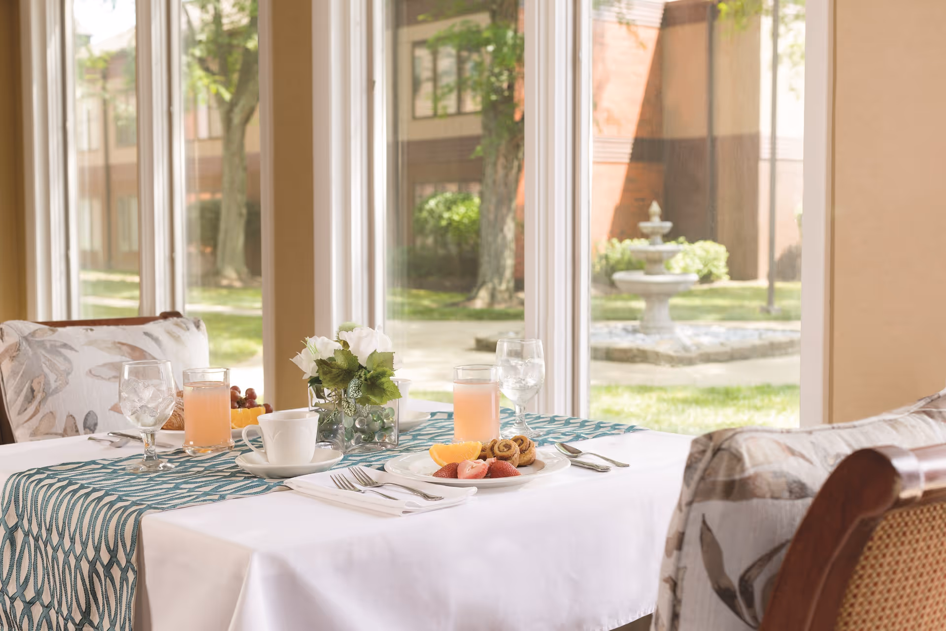 A dining table set for two with a white tablecloth and a blue patterned runner, featuring plates with fruit and pastries, glasses of water and juice, a coffee cup, and a small vase with white flowers. The table is positioned next to large windows overlooking a garden with a stone fountain and trees.