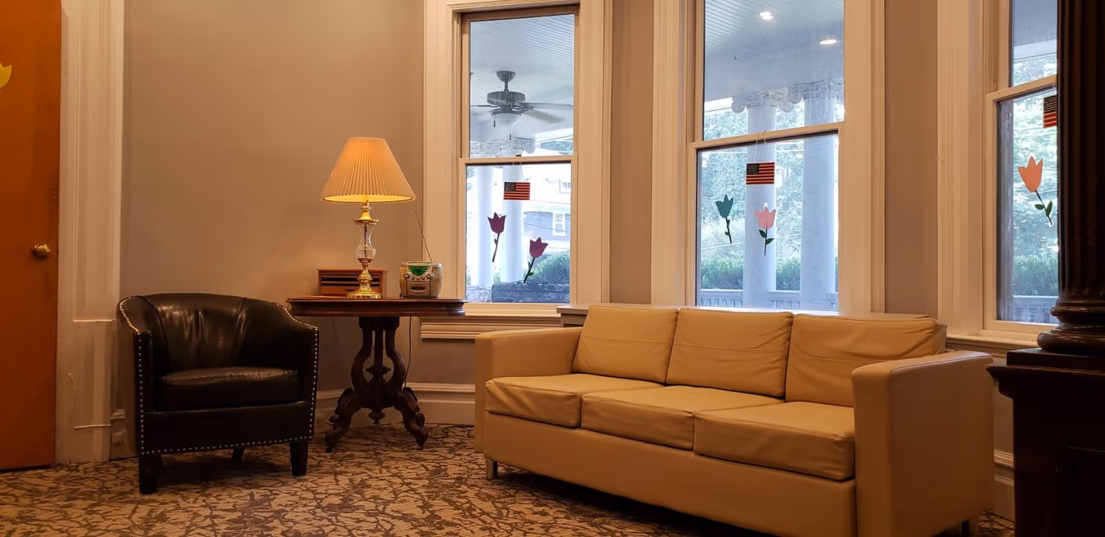 A cozy sitting area in a senior living facility with a beige leather sofa and a black leather armchair. A wooden side table with a lamp and a small radio sits between the chairs. Large windows with decorative flower stickers allow natural light to brighten the room.