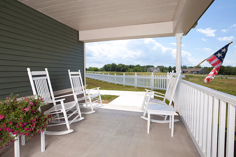 Covered porch with white rocking chairs and a bench, overlooking a grassy yard with a white picket fence and an American flag on the right side. The sky is partly cloudy.