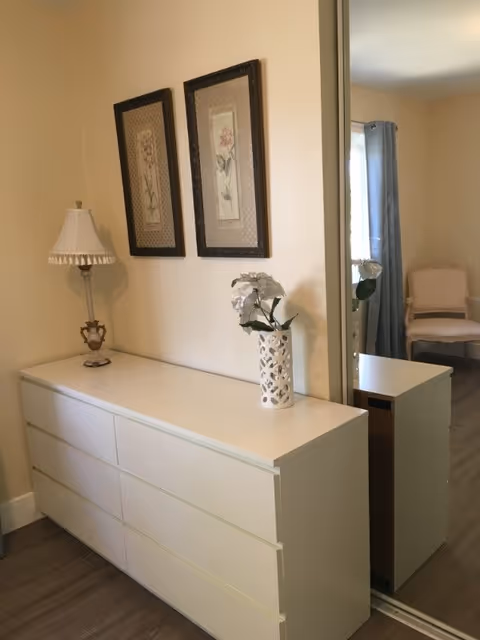 A white dresser with six drawers is placed against a beige wall. On top of the dresser, there is a decorative table lamp with a white lampshade and a white vase holding white flowers. Above the dresser, two framed floral artworks are hung on the wall. To the right, a large mirror reflects part of the room, showing a window with gray curtains and a beige upholstered chair.
