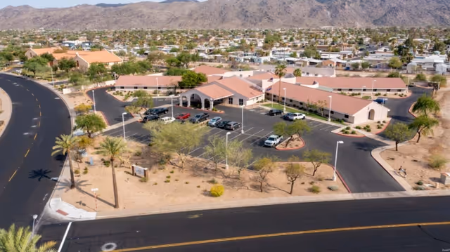 Aerial view of South Mountain Post Acute facility showing a single-story building with a terracotta roof surrounded by parking lots and desert landscaping. The facility is located near a curved road with mountains visible in the background and a residential neighborhood nearby.