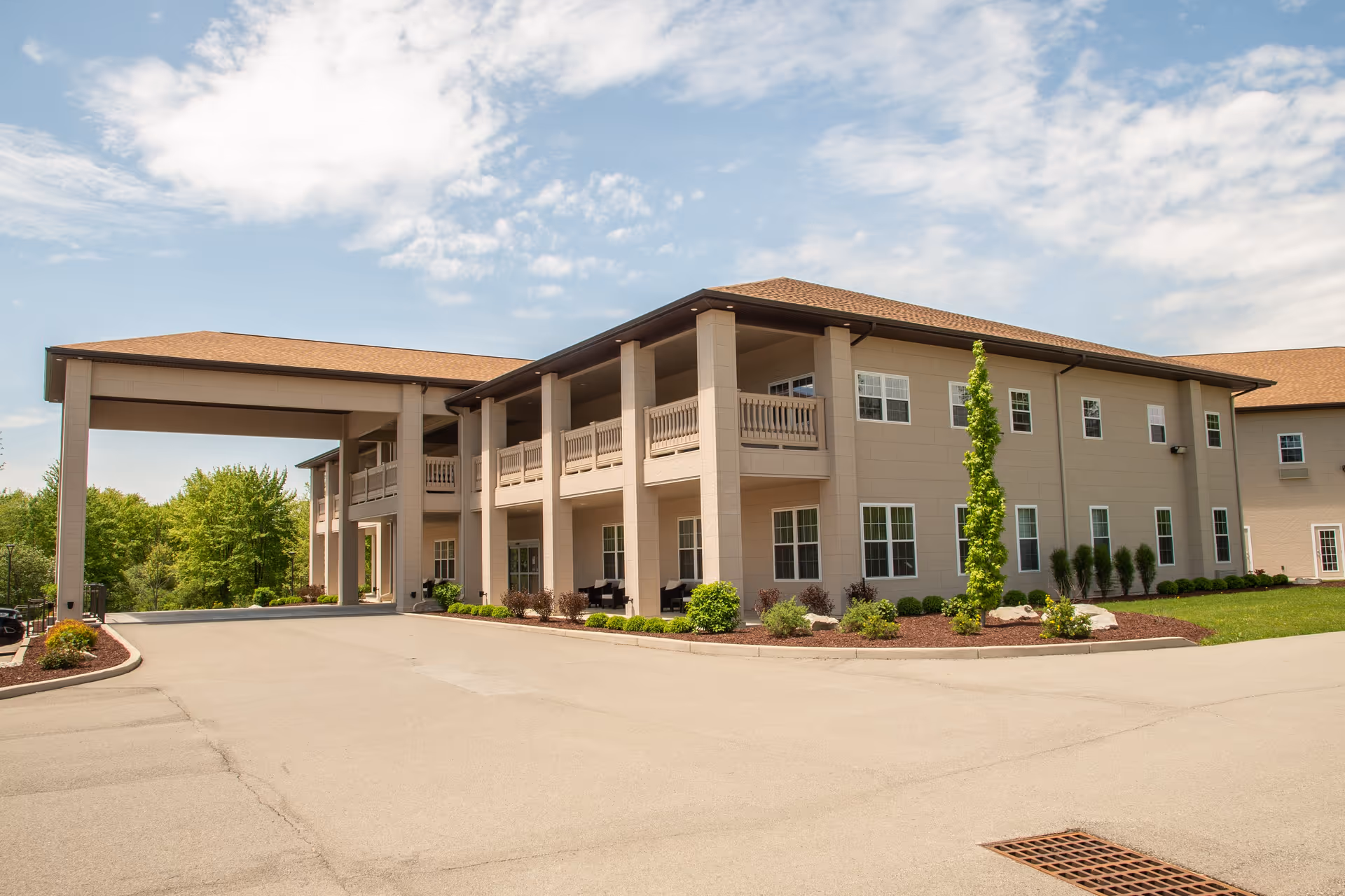Exterior view of a two-story senior living facility building with beige walls and a brown roof. The building features a covered entrance supported by columns, multiple windows, and a landscaped area with small bushes and a young tree. The sky is partly cloudy.
