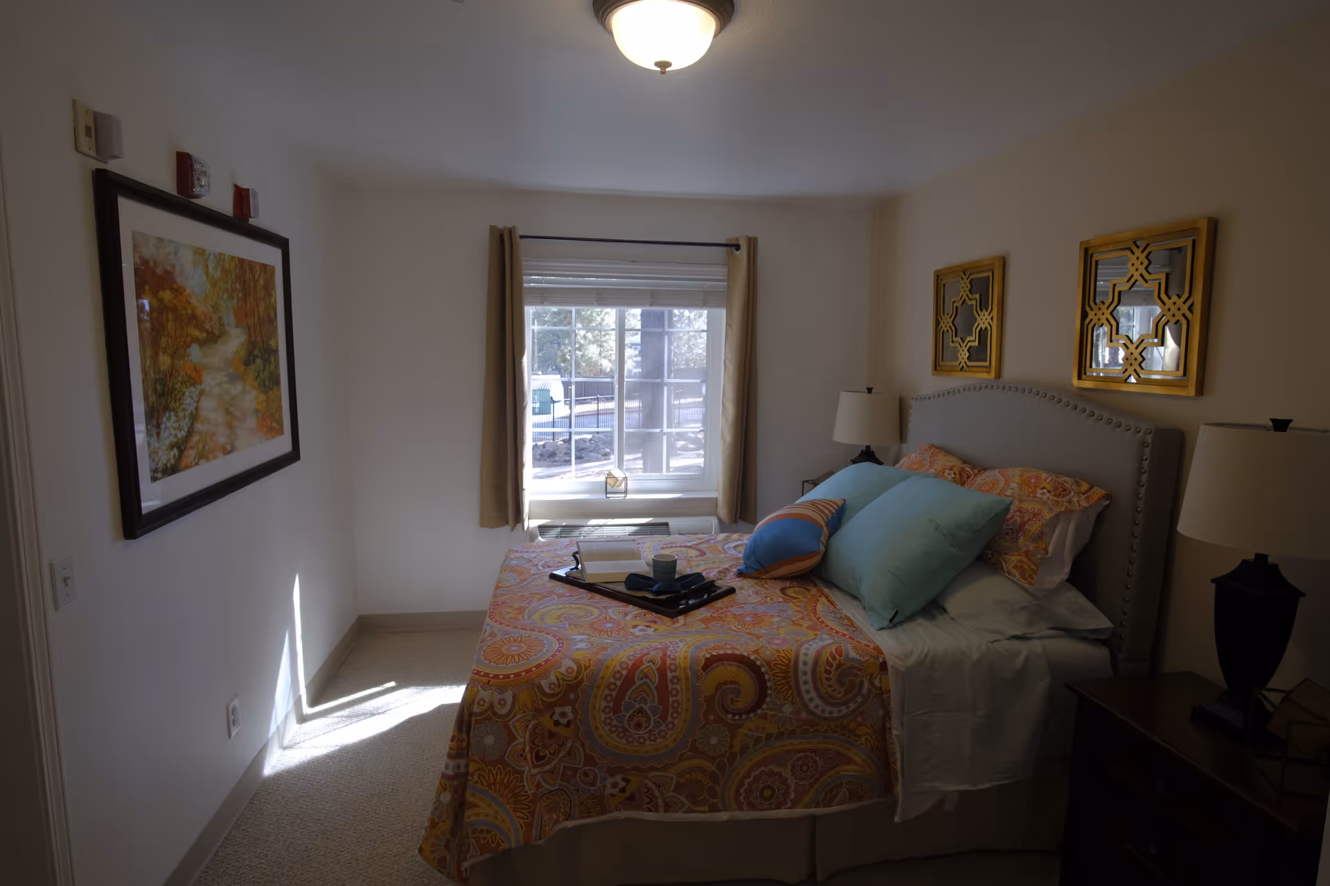 A small bedroom with a bed dressed in a colorful patterned quilt and pillows, nightstands with lamps, framed wall art, and a window letting in daylight.