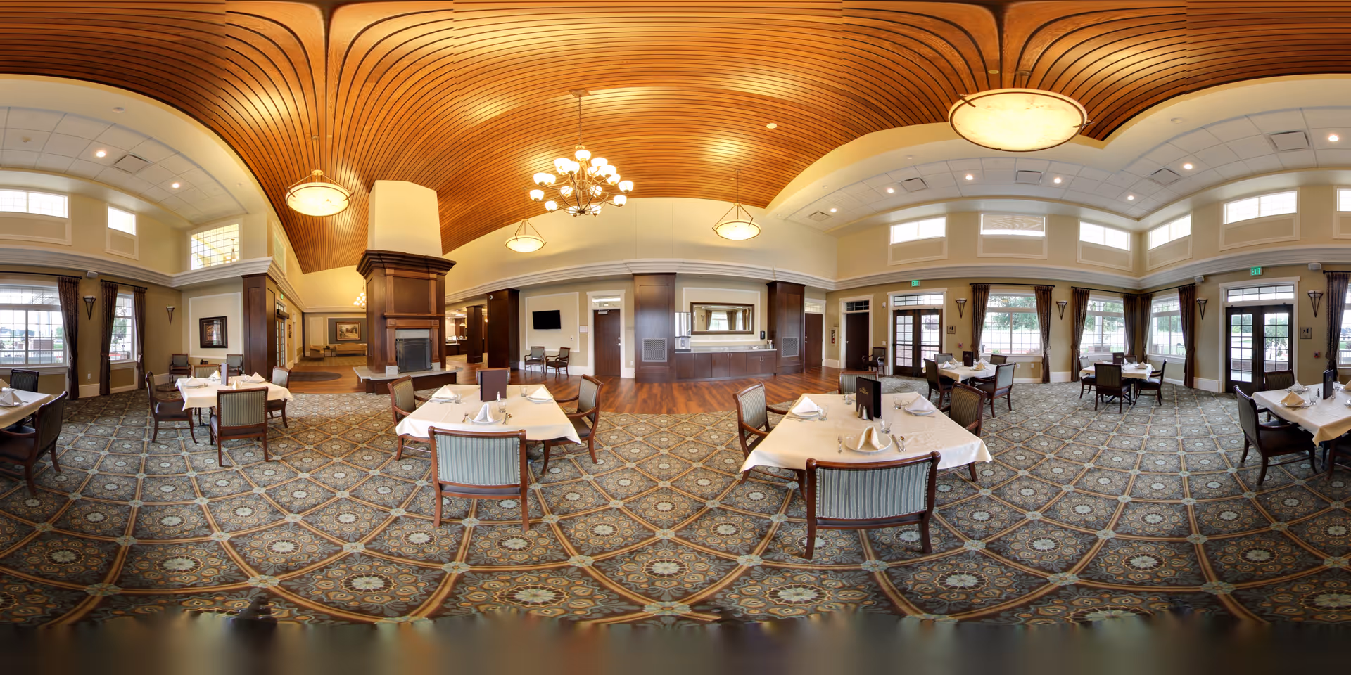 Spacious, elegantly furnished dining room with multiple set tables, patterned carpet, a wood-paneled vaulted ceiling with chandeliers, and large windows.