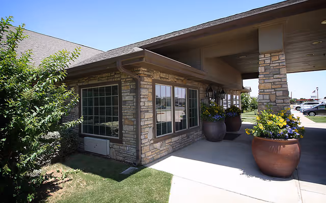 Exterior view of a stone building with large windows and a covered entrance supported by stone pillars. There are large flower pots with colorful flowers near the entrance and a small green bush on the left side. Several parked cars are visible in the background under a clear blue sky.