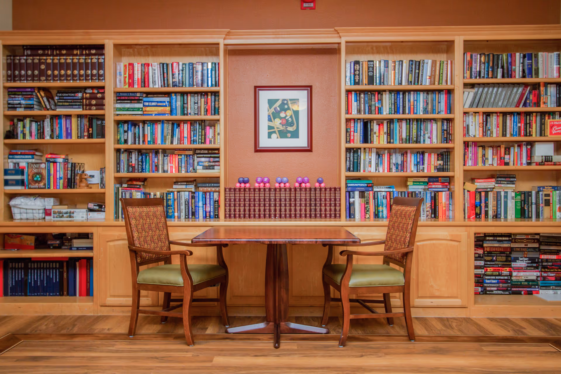 A cozy reading area with a wooden table and two upholstered chairs in front of built-in bookshelves filled with books. A framed picture of a pool table hangs on the wall between the shelves.