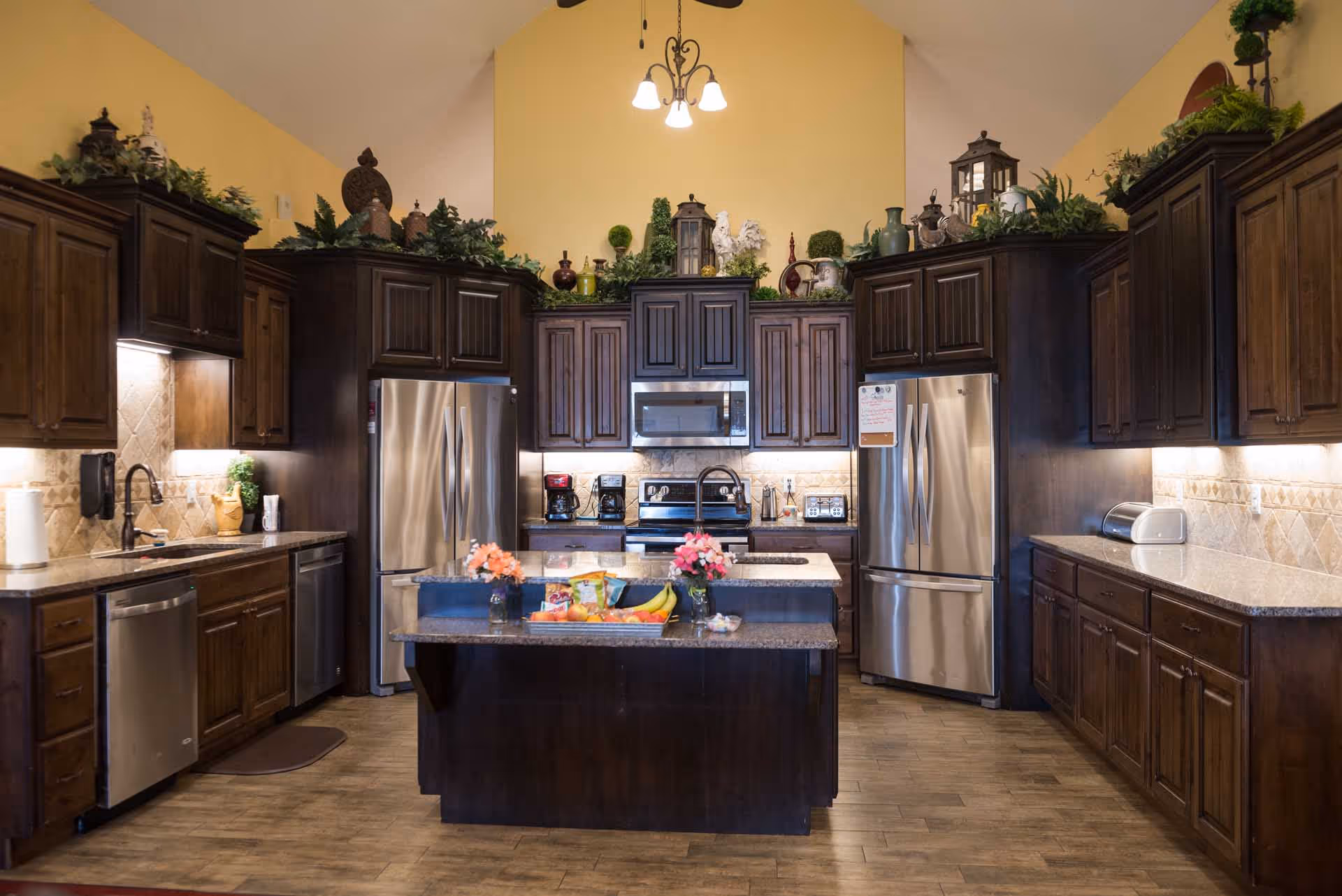 A spacious kitchen with dark wooden cabinets, two stainless steel refrigerators, a microwave, stove, dishwasher, and a central island with flowers and a fruit basket. The kitchen has a high ceiling with a chandelier and decorative greenery on top of the cabinets.