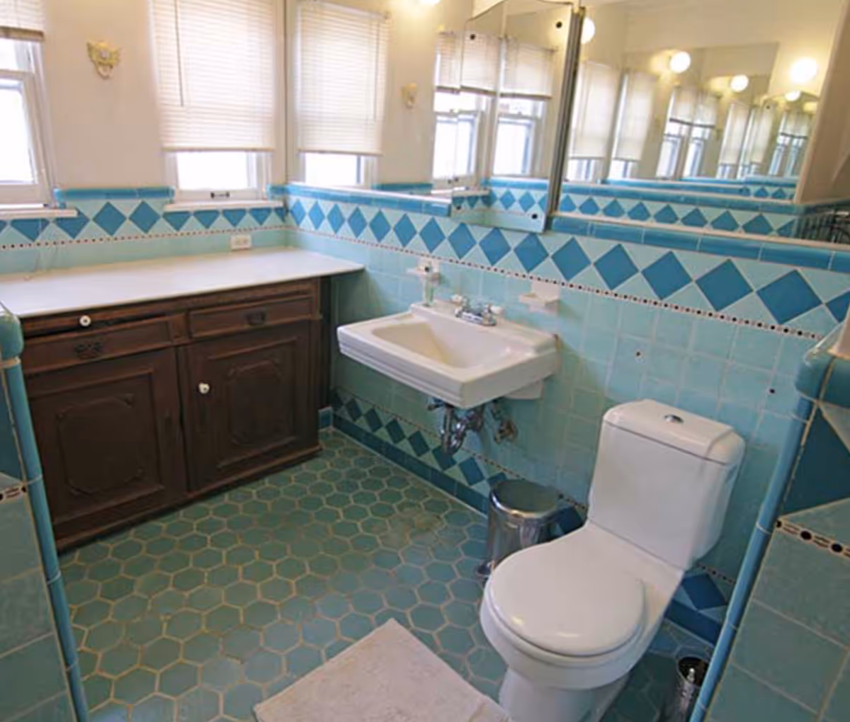 A bathroom with turquoise hexagonal floor tiles and light blue wall tiles featuring a diamond pattern border. The room includes a white toilet, a wall-mounted white sink with a mirror above it, a wooden cabinet with a white countertop, and three windows with blinds partially drawn.