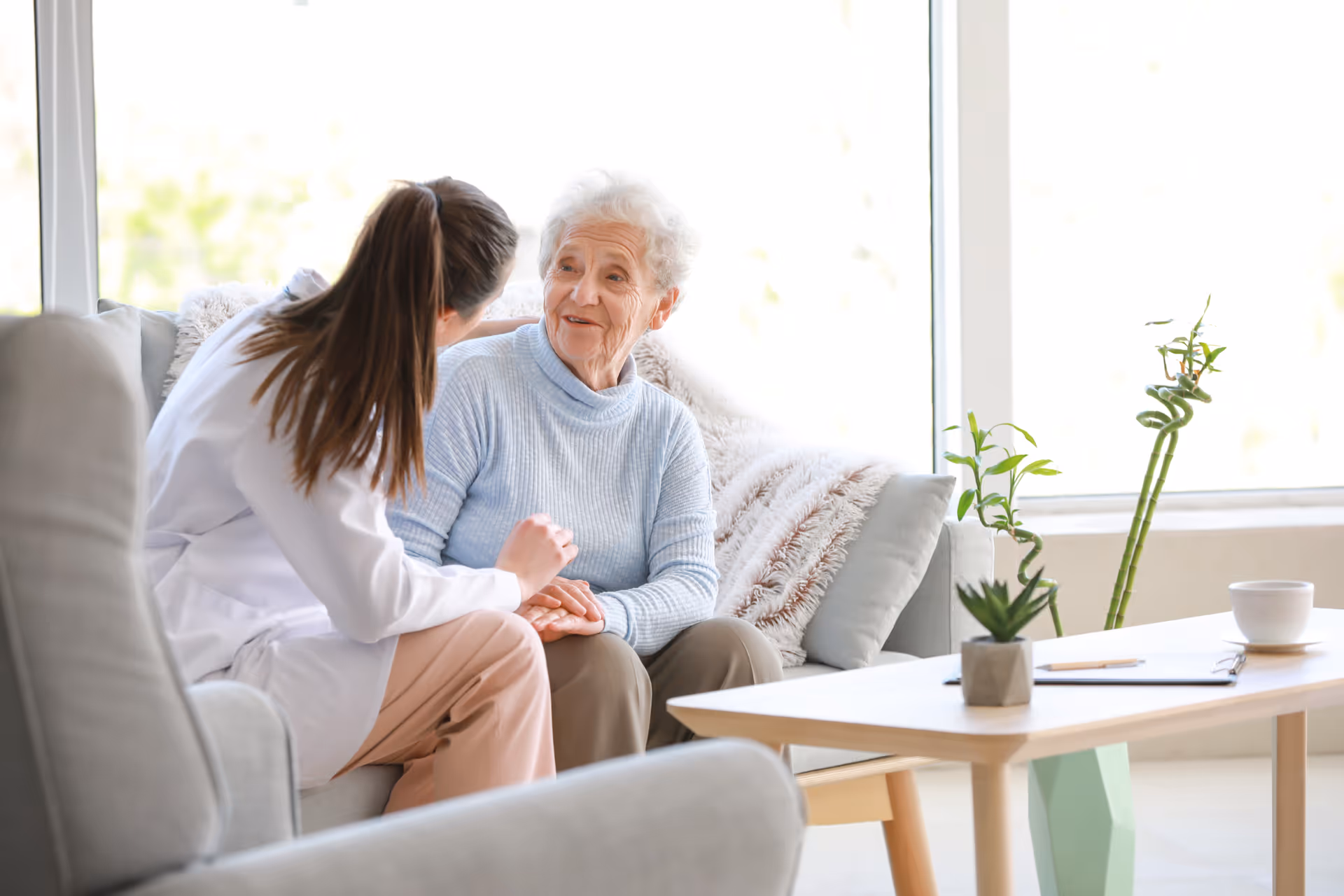 A healthcare professional in a white coat is sitting on a couch and holding hands while talking to an elderly woman with white hair wearing a light blue sweater. They are in a bright room with large windows, a wooden coffee table with plants and a cup, and comfortable seating.