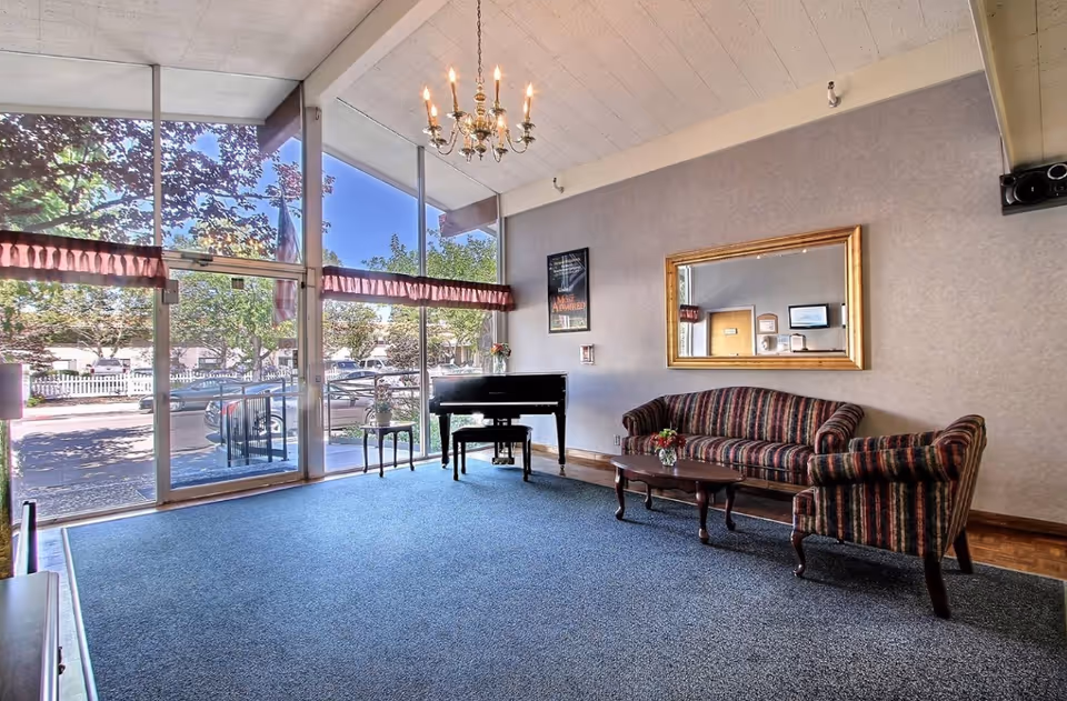 A bright lounge area with large floor-to-ceiling windows showing trees and parked cars outside. Inside, there is a striped sofa and matching armchair with a wooden coffee table in front. A black piano and a small side table with flowers are near the windows. A chandelier hangs from the ceiling, and a large mirror and framed poster are on the wall.