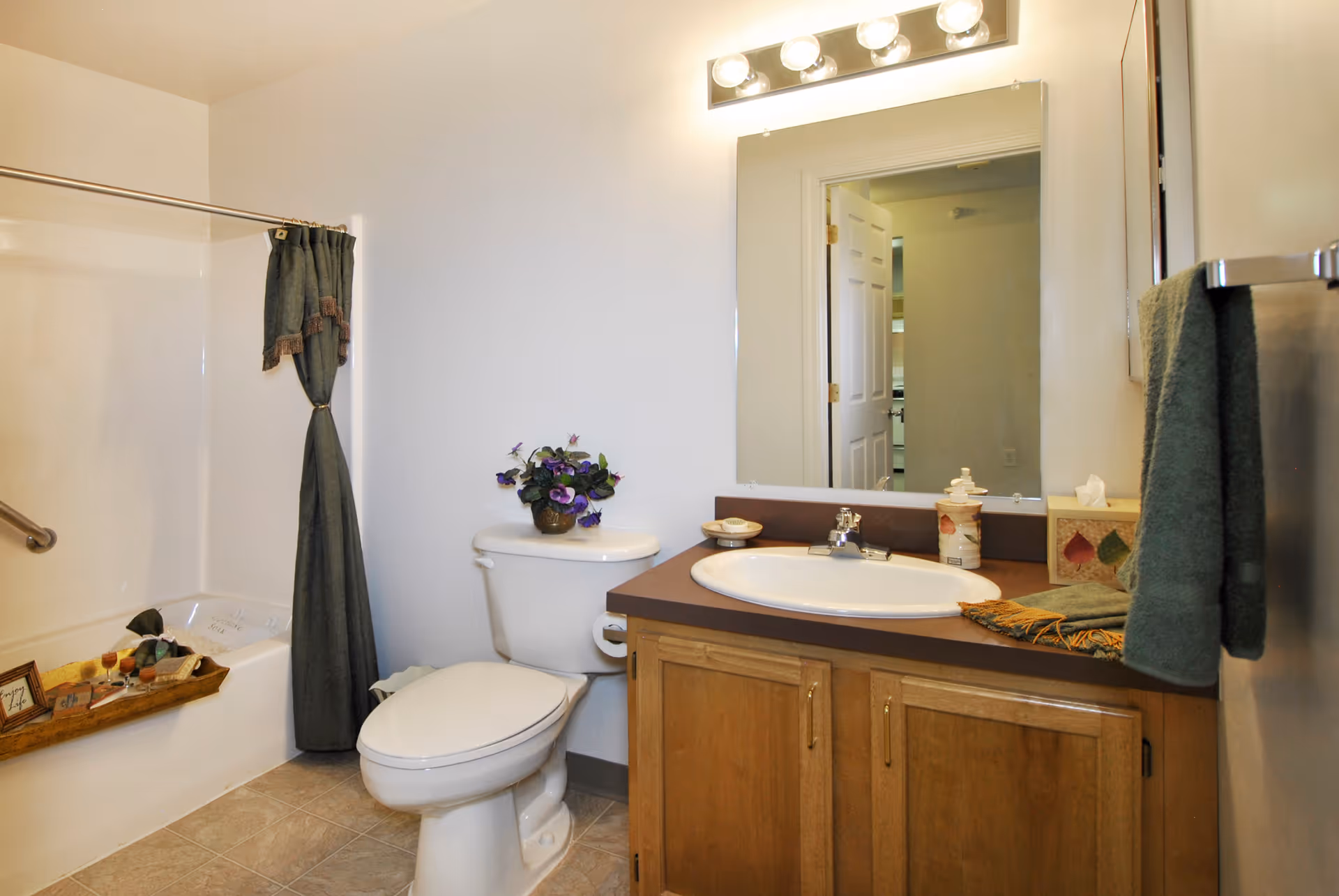 A clean bathroom with a white toilet, a wooden vanity with a sink, a large mirror above the sink, and a bathtub with a dark green shower curtain. There is a towel hanging on a rack and a small decorative plant on the toilet tank.