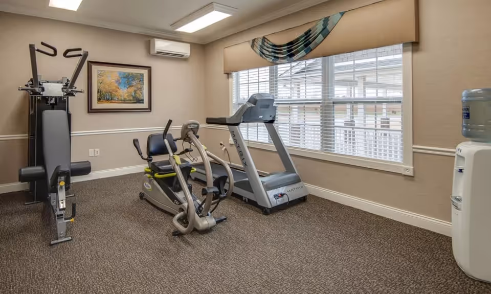 A small fitness room with exercise equipment including a treadmill, a recumbent bike, and a multi-function weight machine. The room has beige walls, a large window with blinds and a decorative valance, a framed landscape painting on the wall, and a water cooler in the corner.