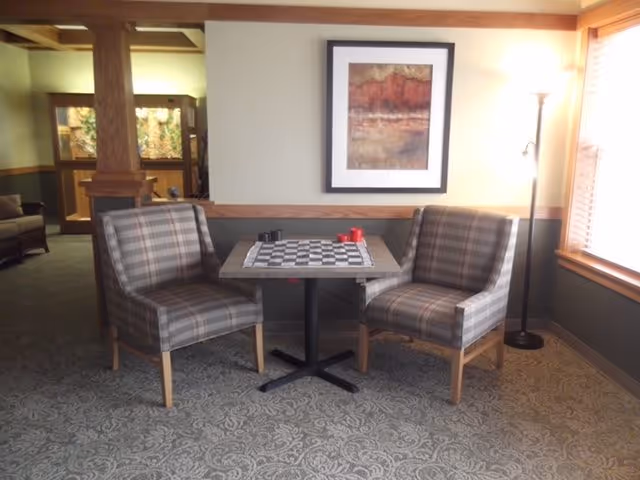 Two plaid upholstered chairs face a small table with a checkerboard in a carpeted seating area with framed art and a floor lamp.