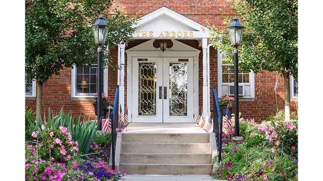 Entrance to a brick building with white double doors featuring decorative glass panels. The entrance is framed by a white portico with the words 'THE ARBORS' above the doors. There are steps leading up to the doors, flanked by black railings and two black lamp posts. The area around the entrance is landscaped with green shrubs, pink and purple flowers, and small American flags.