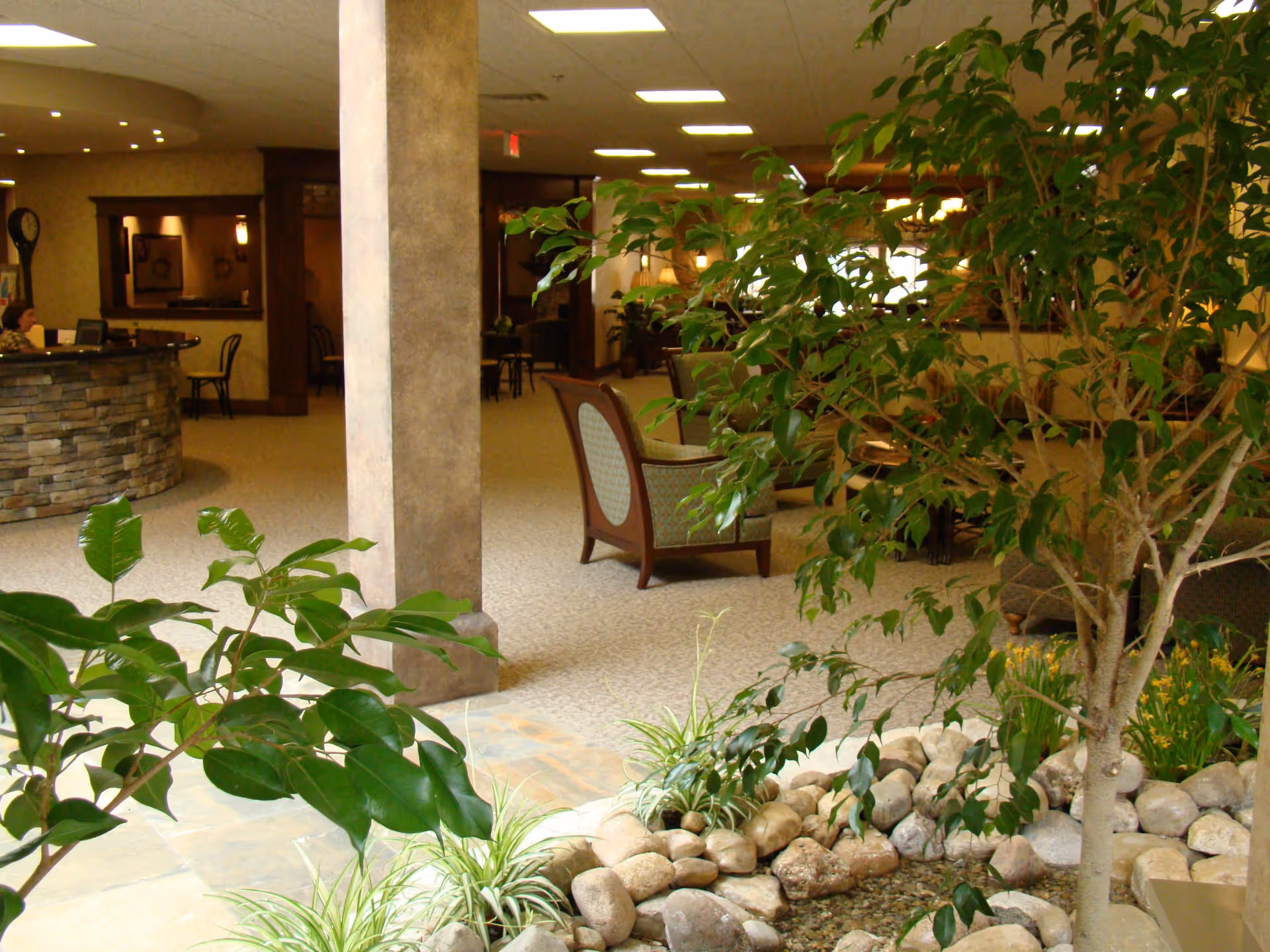 Indoor senior living lobby with seating, a stone reception desk, indoor plants and a rock-lined water feature.
