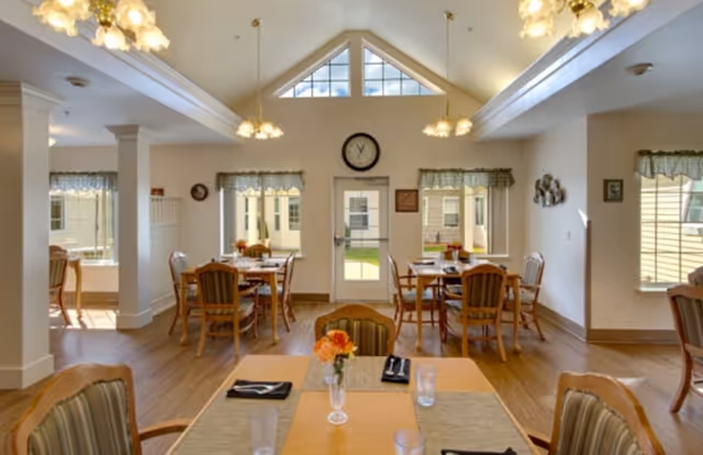 A bright dining room with wooden tables and chairs arranged neatly. Each table has a small flower vase with orange flowers and place settings including glasses and napkins. Large windows with valances let in natural light, and a clock is mounted above a door at the far end of the room. The ceiling is vaulted with hanging light fixtures.