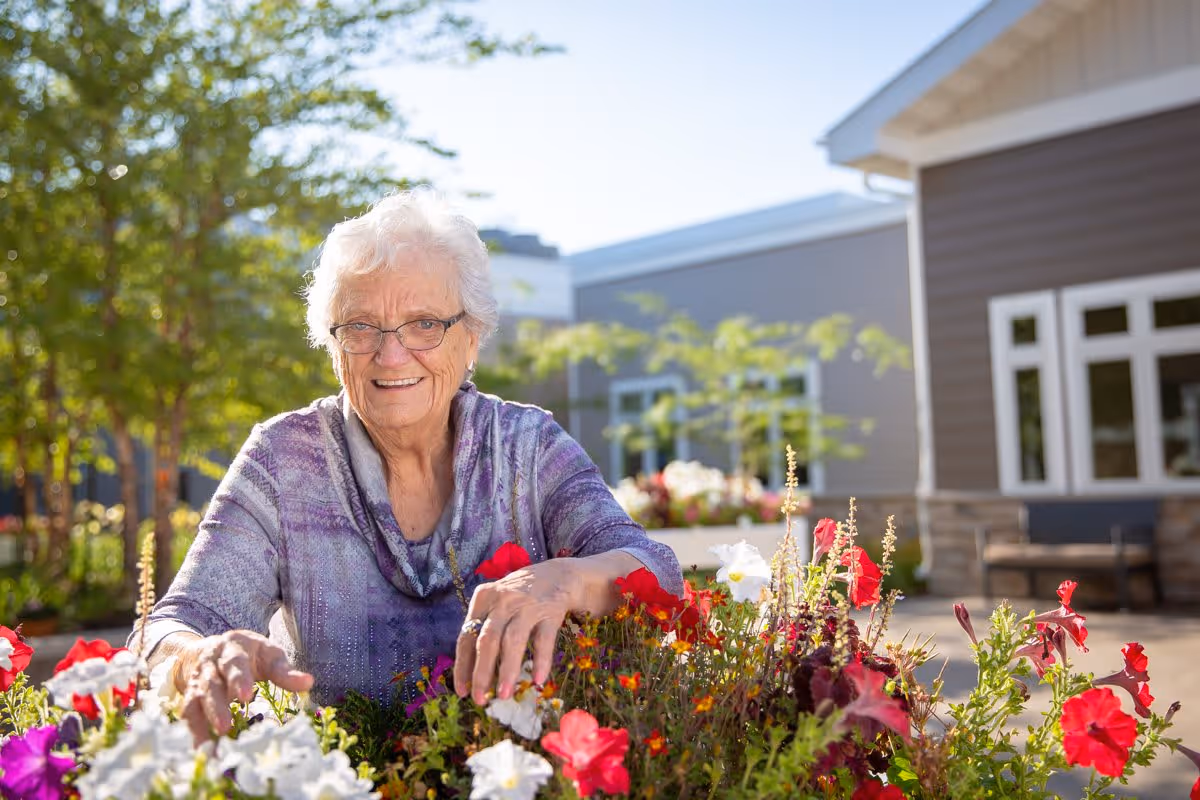 An elderly woman with white hair and glasses wearing a purple patterned top is smiling while tending to a colorful flower bed outdoors. Behind her, there is a building with large windows and a bench, and trees with green leaves are visible in the background under a clear sky.