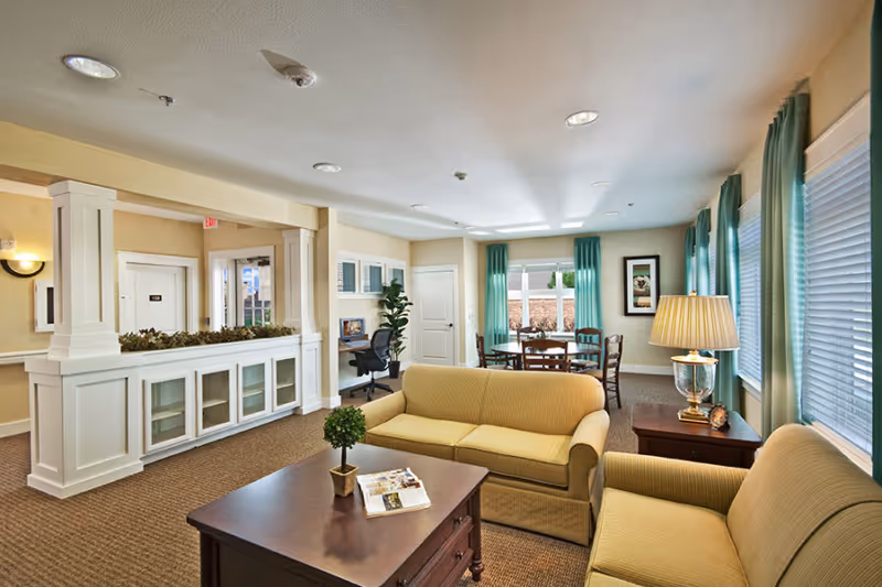 A bright and spacious living room area in a senior living facility with beige sofas, a wooden coffee table with a small potted plant and magazines, a side table with a lamp, and a round dining table with chairs near windows with teal curtains. There is also a small desk with a computer and chair against the wall.