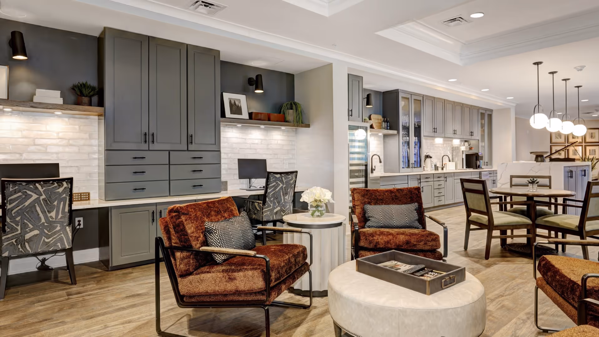 A modern senior living common area featuring two rust-colored armchairs with patterned pillows, a round ottoman with a tray, a small round side table with a flower vase, and a dining table with four chairs. The background includes a long counter with gray cabinets, a built-in desk area with chairs and computers, and pendant lights hanging from the ceiling.