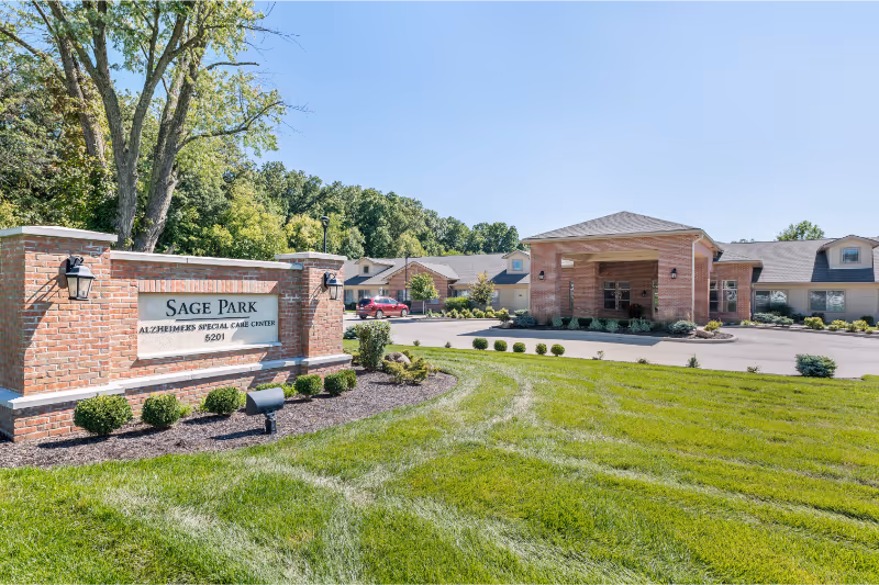 Exterior view of Sage Park Alzheimer's Special Care Center showing a brick sign with the facility name and address, a well-maintained lawn, and a large building with a covered entrance under a clear blue sky.