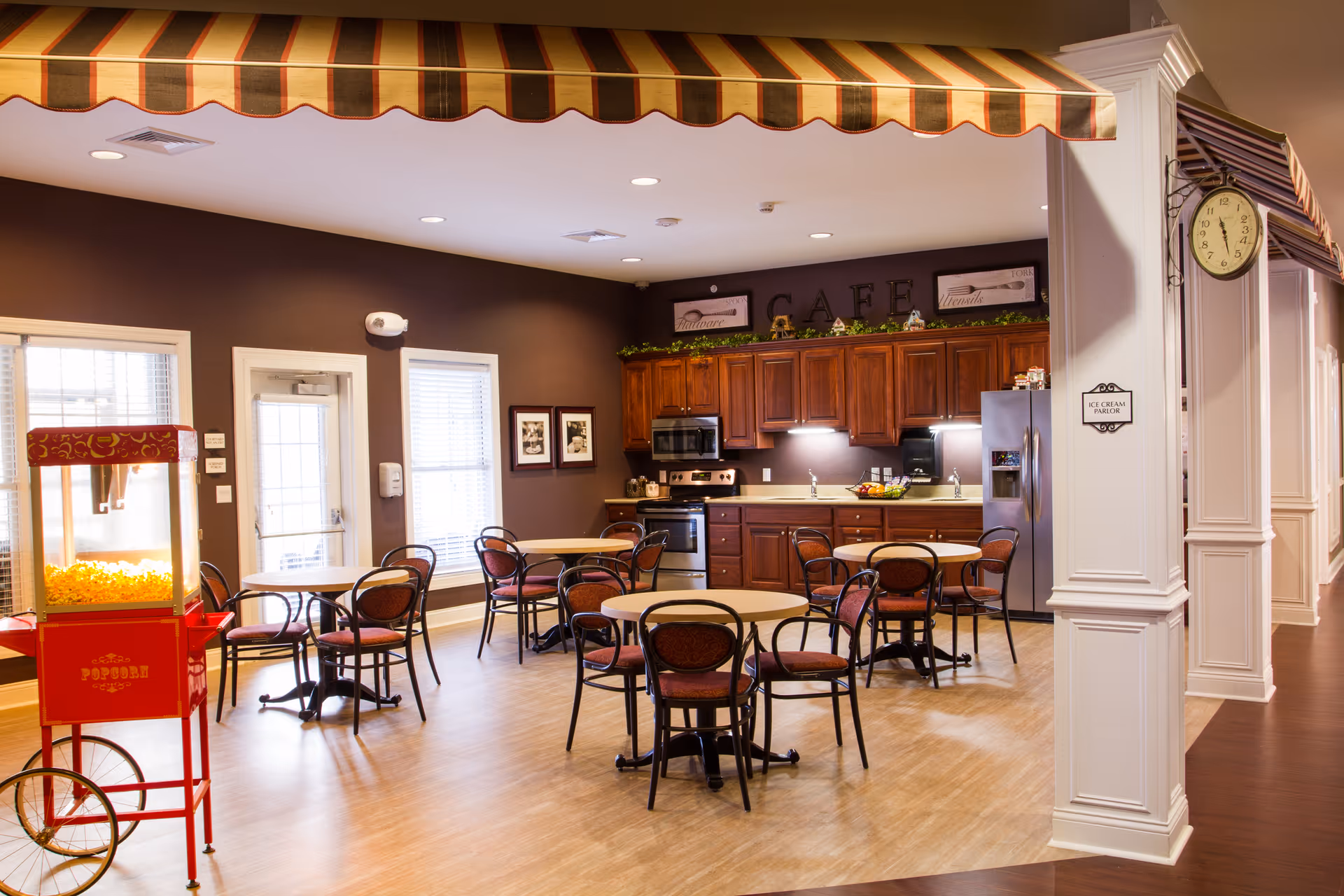 Communal dining area with round tables and chairs, a popcorn cart, and a kitchen/café counter under a striped awning.