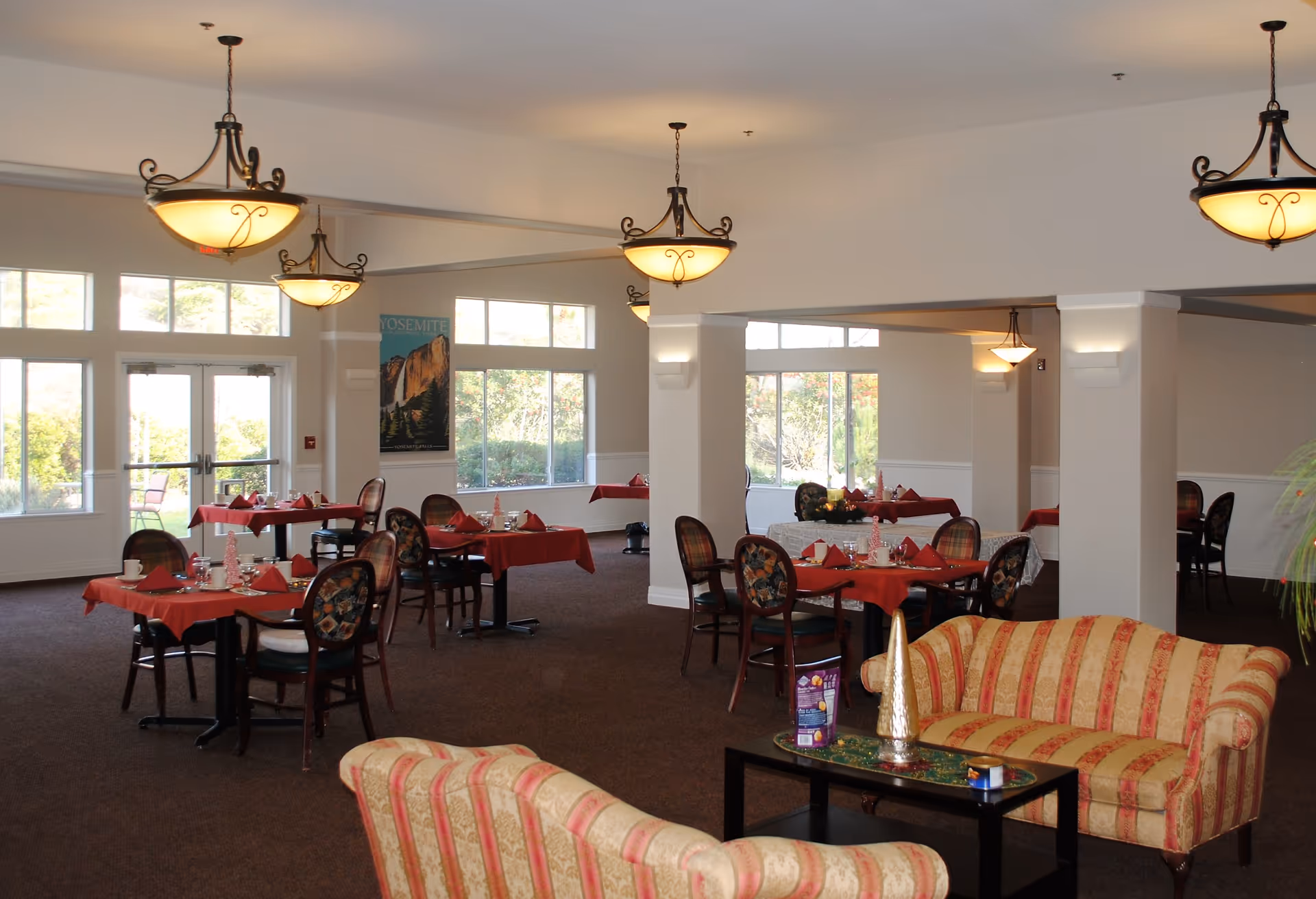 Communal dining room with multiple red-tablecloth tables, chandeliers, large windows, and a seating area with striped couches.