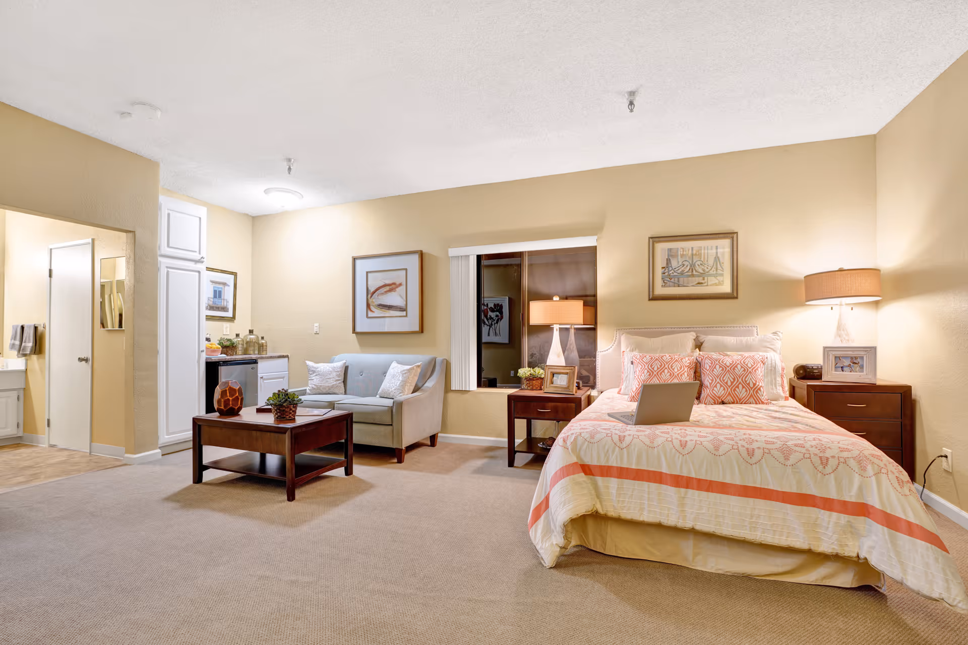 A cozy senior living facility room at The Montera featuring a bed with patterned bedding and pillows, two bedside tables with lamps and framed photos, a small sofa with cushions, a wooden coffee table with decorative items, a kitchenette area with a mini fridge and cabinets, and an open doorway leading to a bathroom.
