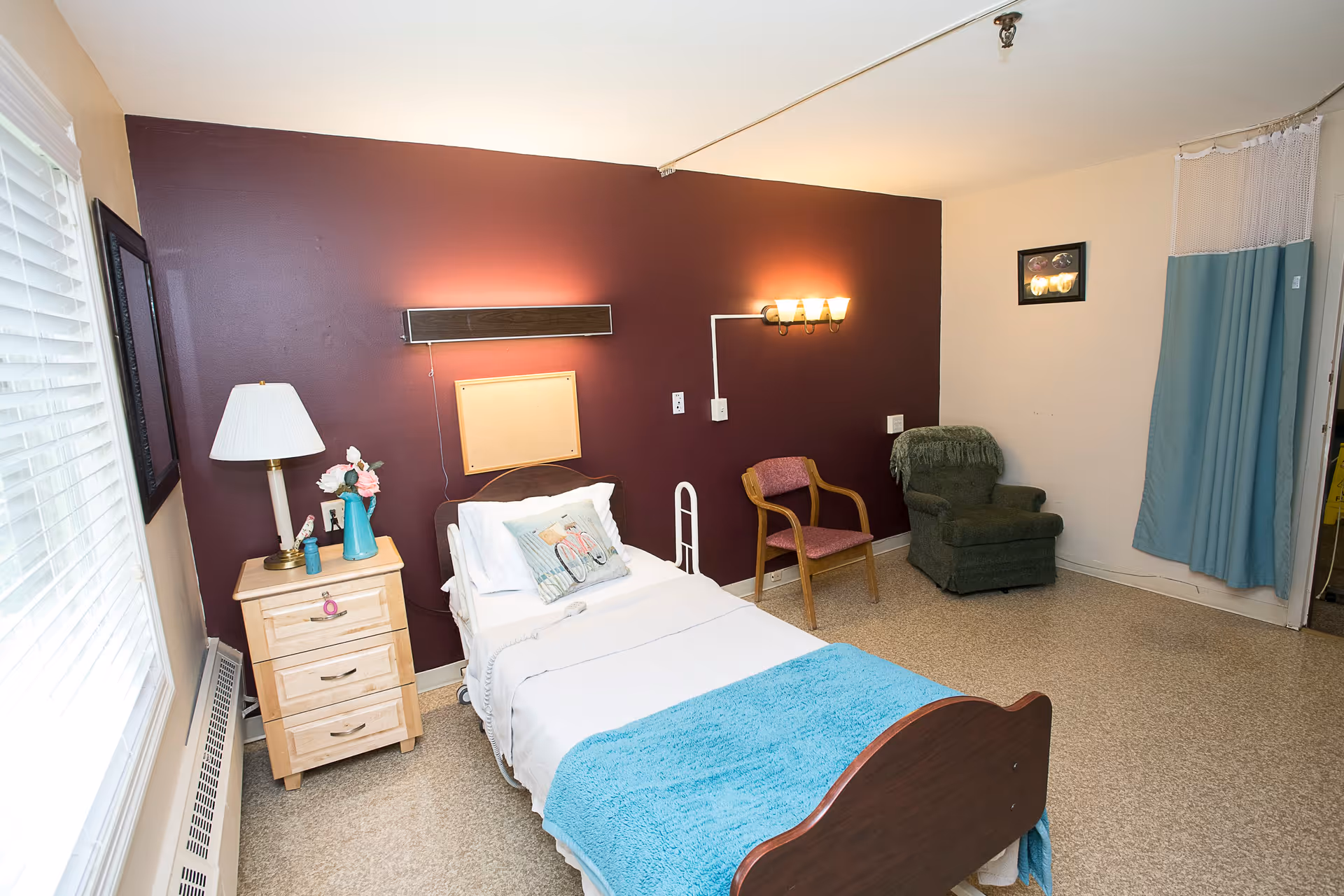 A small senior care bedroom with a single bed, bedside table and lamp, seating chairs and a privacy curtain.