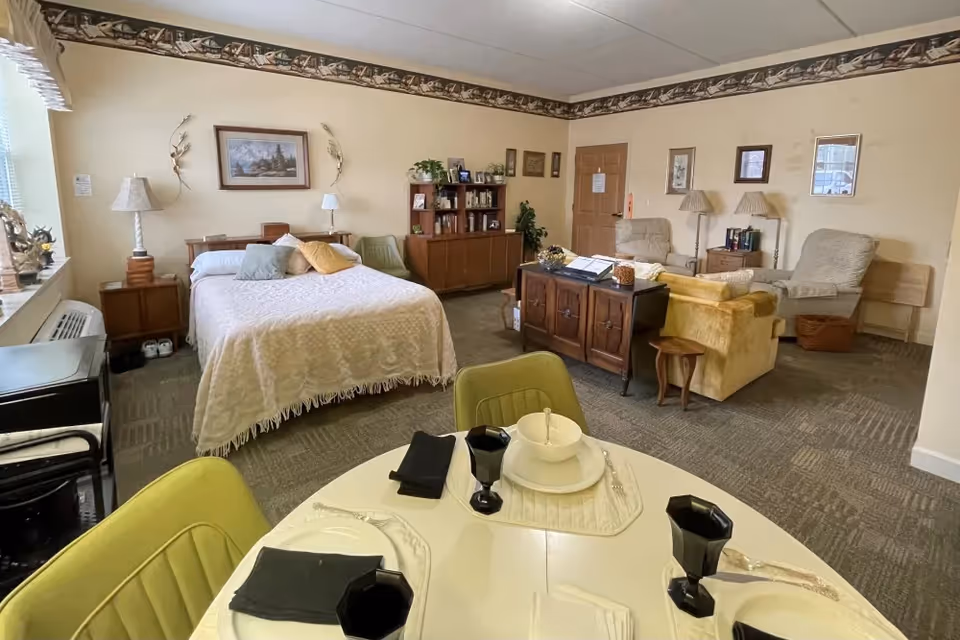 A cozy, well-lit room featuring a bed with pillows and a white bedspread, a wooden bookshelf, two armchairs, a yellow sofa, and a small dining table set with plates, bowls, black glasses, and napkins. The room has beige walls with framed pictures and decorative wall trim near the ceiling.