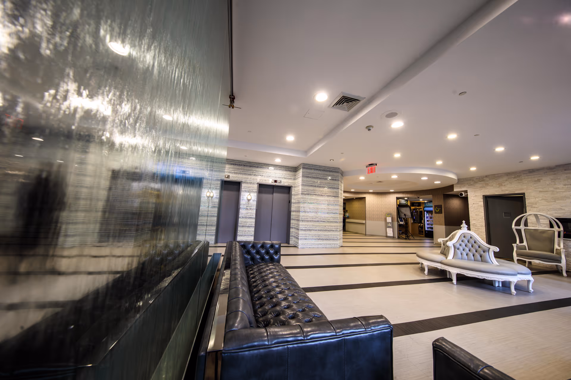 Modern and spacious lobby area with black leather tufted sofas, a white ornate chaise lounge, and a white chair with a rounded back. The floor has a striped pattern, and there are two elevators with a textured wall behind them. The ceiling has recessed lighting and a circular architectural feature. A water feature or reflective glass wall is visible on the left side.