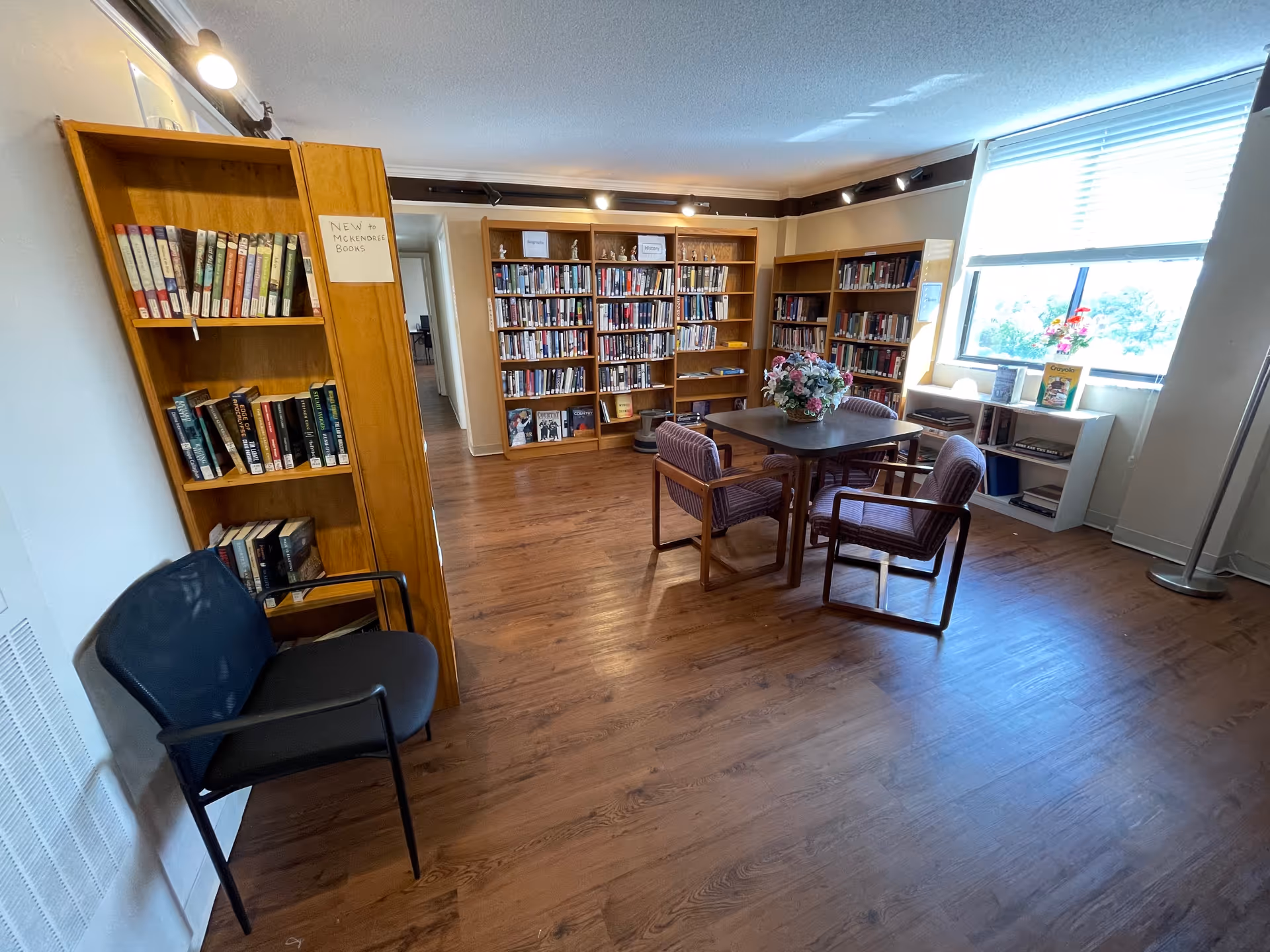 A cozy library room with wooden bookshelves filled with books along the walls. There is a square table in the center with four cushioned chairs around it and a flower arrangement on top. A window on the right side lets in natural light, and a black chair is positioned near a bookshelf labeled 'New to McKendree Books'. The room has wooden flooring and soft lighting.