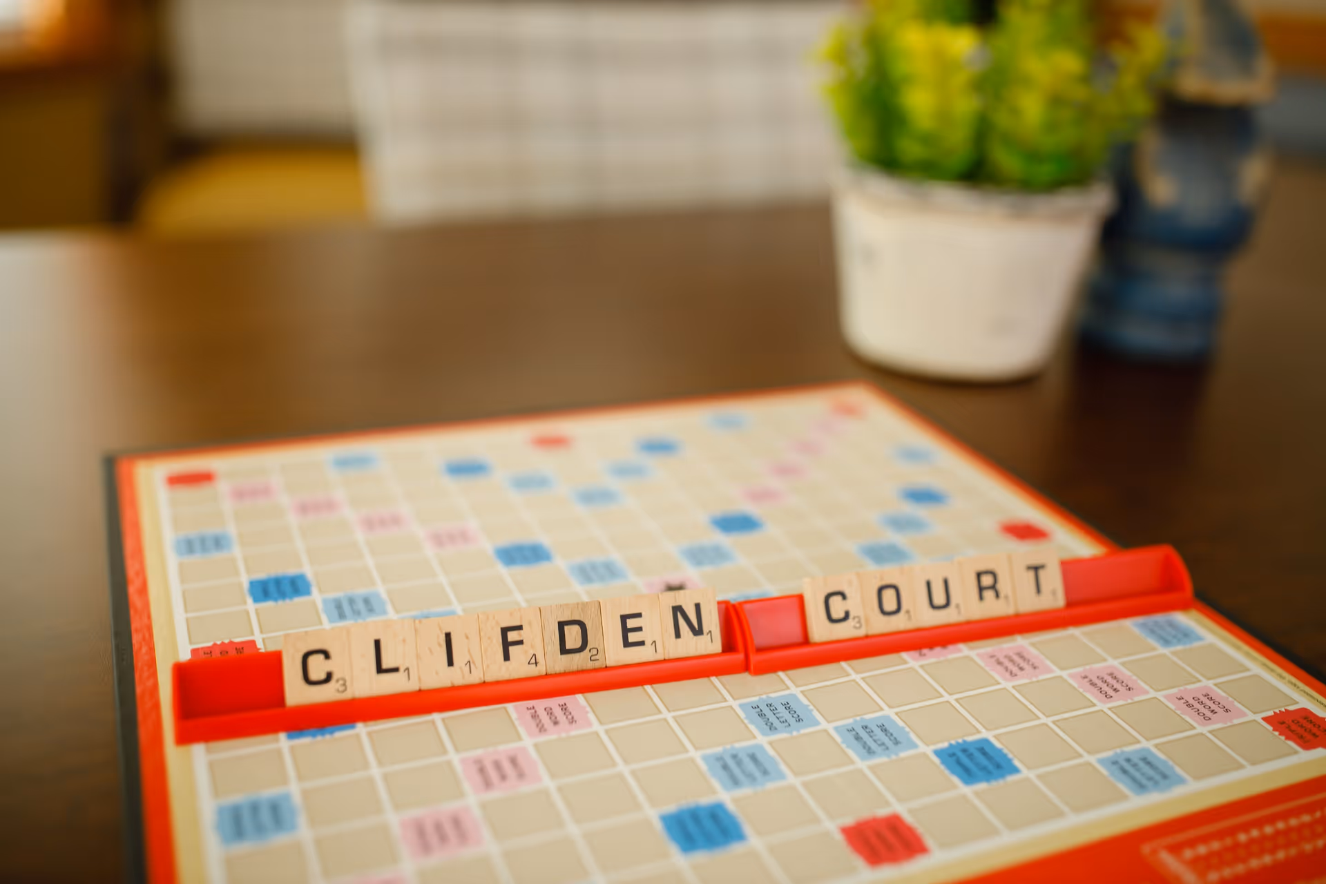 A close-up view of a Scrabble game board on a wooden table with letter tiles arranged to spell 'CLIFDEN COURT' on a red tile rack. In the background, there is a blurred white pot with green plants and a blue decorative object.