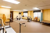 A spacious physical therapy or rehabilitation room with exercise equipment including stationary bikes, parallel bars for walking practice, a treatment table, and a chair near the window. The room has beige walls, carpeted floor, and fluorescent ceiling lights.