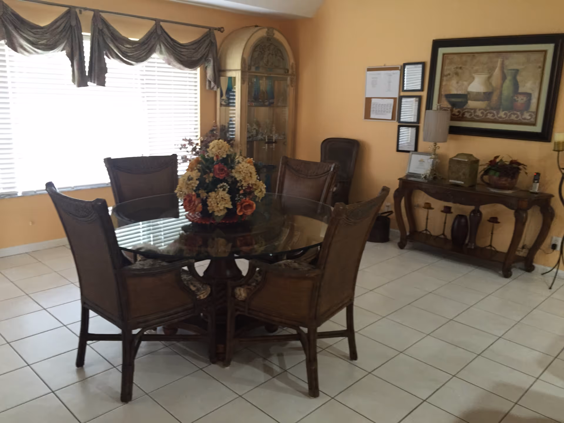 A dining area with a round glass-top table surrounded by six wicker chairs. A floral centerpiece is placed on the table. The room has light-colored tiled flooring and peach-colored walls. There is a large window with blinds and decorative curtains on the left side. In the background, there is a wooden display cabinet with glass doors, a wooden console table with decorative items, a lamp, and framed artwork on the wall.