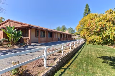 Single-story building with a covered porch, white split-rail fence, and a manicured lawn under a clear blue sky.