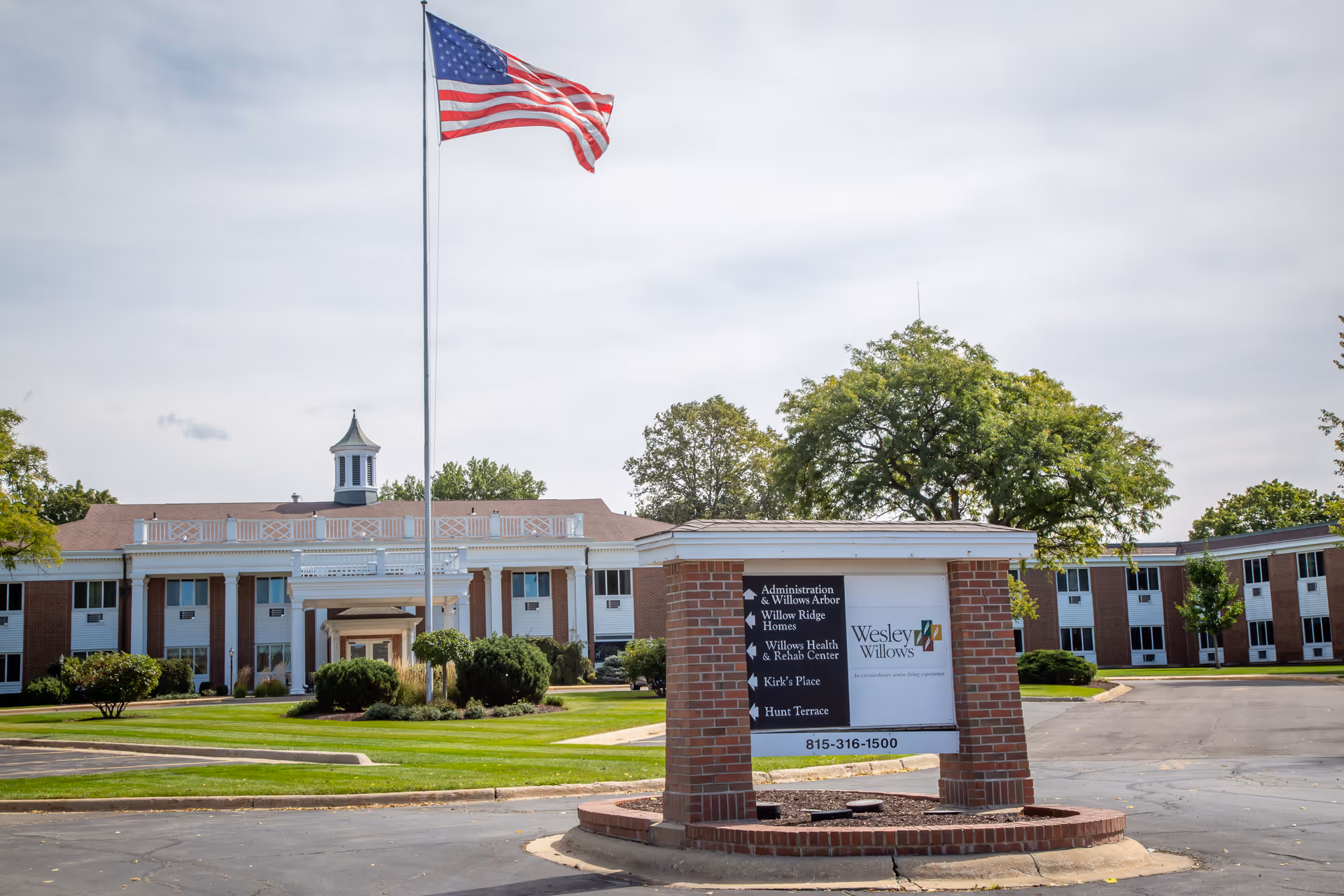 Exterior view of Wesley Willows senior living facility with a large American flag on a flagpole in front. The building is a two-story structure with white columns and brick accents, surrounded by green lawns and trees. A sign in front displays directions to various parts of the facility and the Wesley Willows logo.