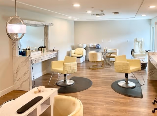 Interior view of a senior living facility's salon area with cream-colored salon chairs, a large mirror with a marble frame, hair care products on the counter, and a wooden floor. The room is well-lit with ceiling lights and has a clean, modern design.