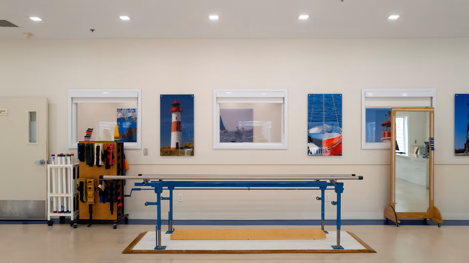 A physical therapy or rehabilitation room with parallel bars for walking exercises, a rack holding various exercise bands and equipment, a full-length mirror on wheels, and framed pictures of lighthouses and boats on the wall.