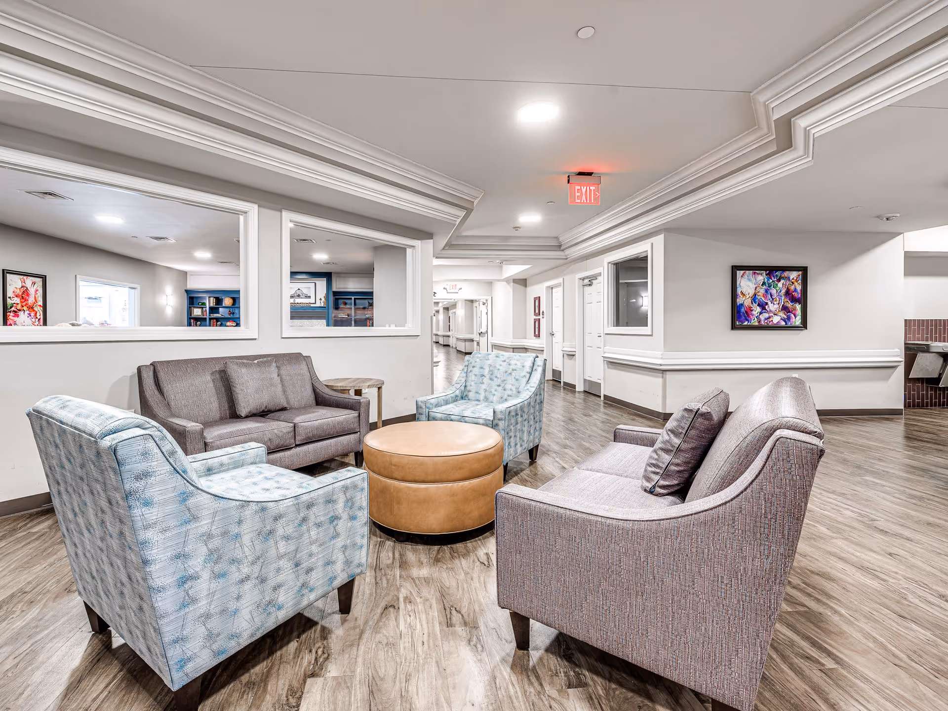 A bright and spacious seating area in a senior living facility featuring two gray sofas, two light blue patterned armchairs, and a round tan ottoman in the center. The room has wood flooring, white walls with crown molding, and colorful abstract artwork on the walls. There are windows looking into an adjacent room and a hallway visible in the background.