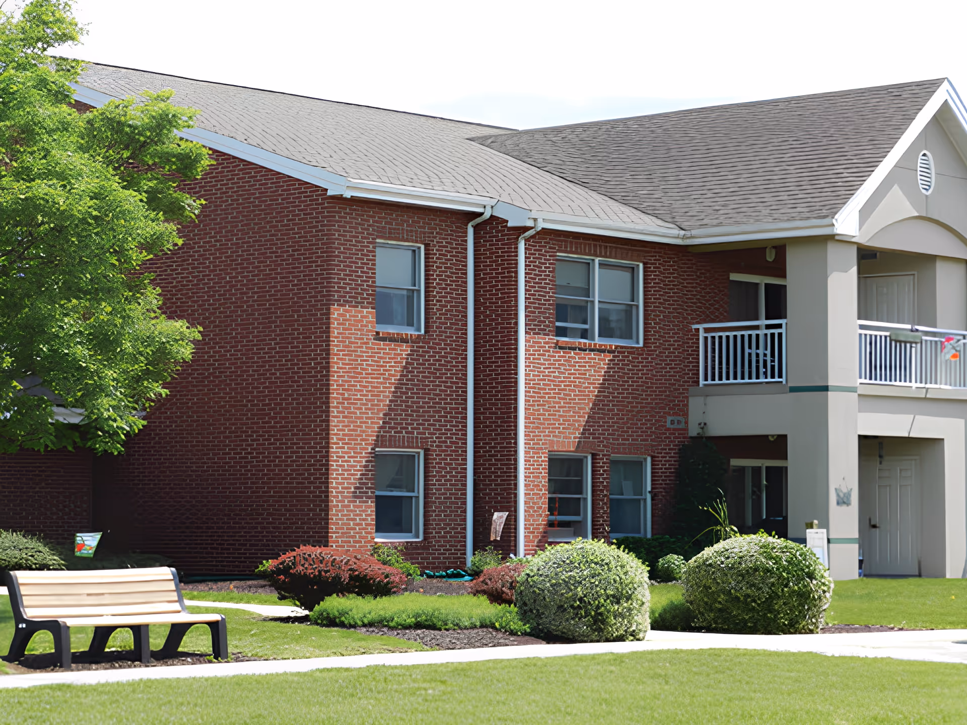 Exterior view of a brick building with multiple windows, a small balcony with white railing, and a well-maintained lawn with bushes and a wooden bench on a concrete path.